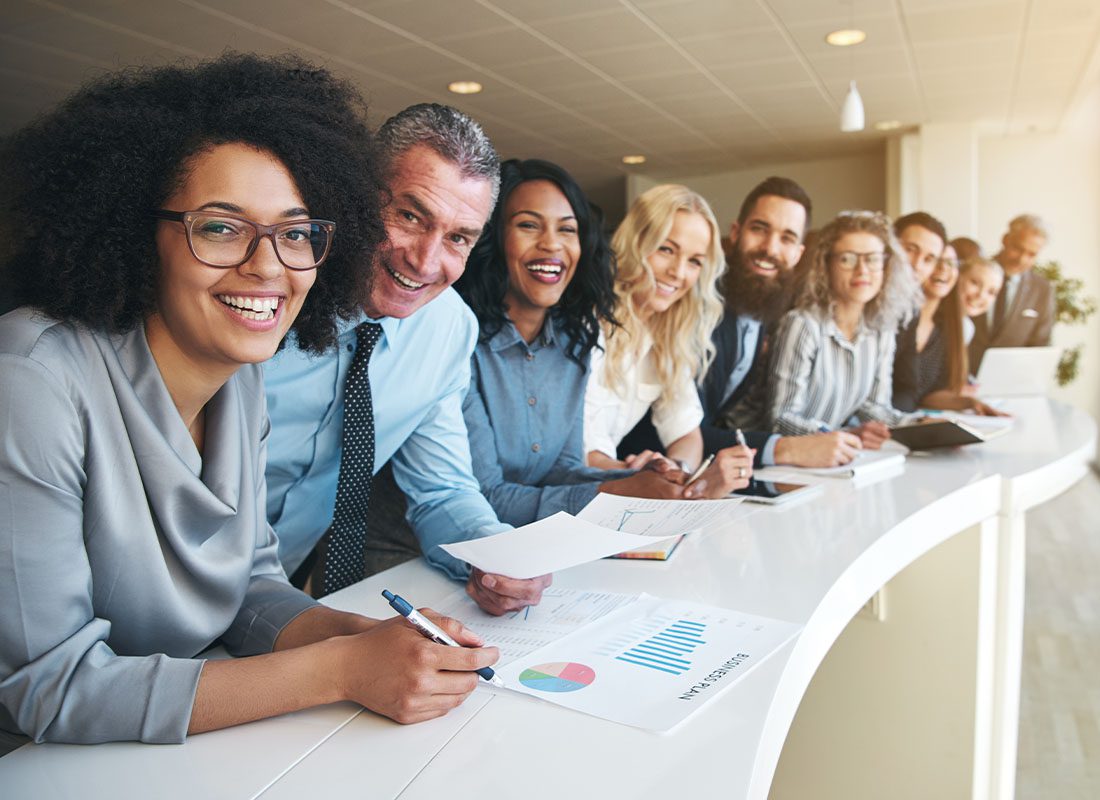 About Our Agency - Cheerful Colleagues Forming a Line and Leaning Against a Desk Looking at Camera in Office